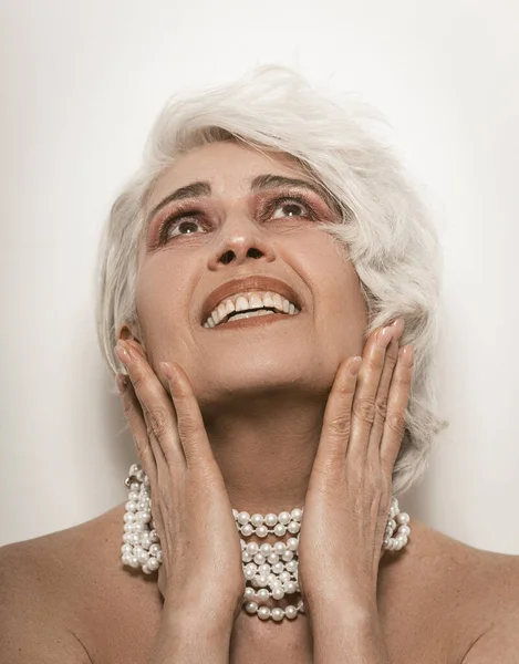 Naken äldre kvinna toothy leende röra hennes ansikte med händerna. Naken mogen kvinna med pärlpärlor runt halsen på vit backgroumd. Vård för mogen hud koncept. Tonad bild — Stockfoto