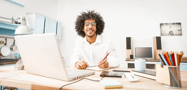 Усміхнений бізнесмен працює ноутбуком у себе вдома. Нахабний арабський хлопець у окулярах ловить фотоапарат, сидячи за столом. Концепція фрілансу. Робота з домашньою концепцією — стокове фото