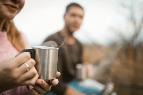 Két turista iszik egy forró italt a természetben. Szelektív összpontosítani fém kemping csésze gőz forró kávé vagy tea női kezekben. Kaukázusi fickó mosolygós lánnyal pihenjen a szabadban. Közelkép. — Stock Fotó