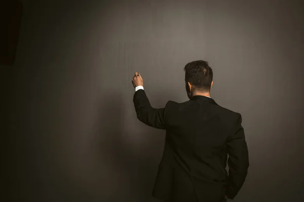 Rückansicht des Geschäftsmannes im schwarzen Anzug schreibt etwas an die dunkelgraue Wand. Seriöser kaukasischer Mann in Formalbekleidung, der dem Betrachter auf grauem Hintergrund den Rücken zuwendet — Stockfoto