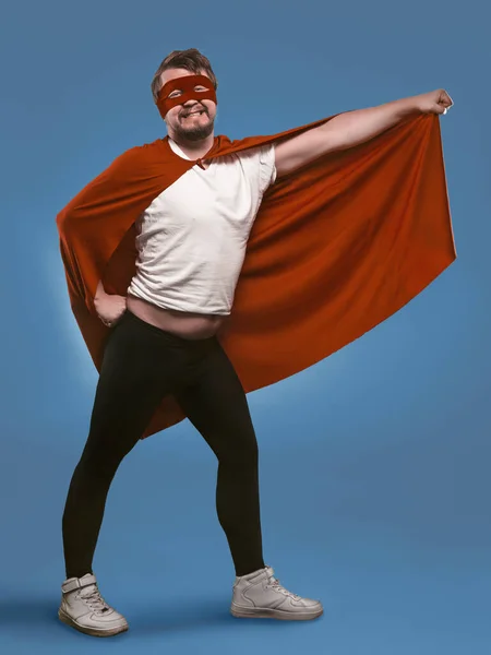 Sorrindo super herói homem pronto para voar salvar o mundo. Homem engraçado em traje de super-herói vermelho e máscara segurando seu manto com a mão posando em pleno crescimento no fundo azul denim desbotada — Fotografia de Stock