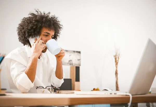 Il giovane uomo d'affari parla sul cellulare mentre lavora con un computer. Arabo ragazzo beve caffè mentre seduto a una scrivania in ufficio o interno della casa — Foto Stock