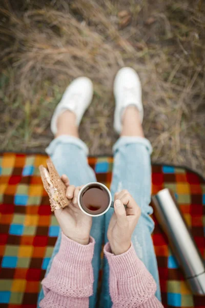 Picknick oder Snack in der Natur. Weibliche Hände halten eine Tasse Tee und ein Sandwich in der Hand. Junge Frau oder Teenie-Mädchen naschen, während sie auf Gras sitzen. Nahaufnahme. Blick aus dem Hochwinkel — Stockfoto