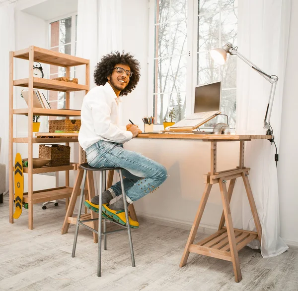 Smart millennial portátil de trabajo en casa sentado cerca de la ventana. Árabe freelancer en camisa blanca y jeans sonríe mirando a la cámara. Trabajo desde el concepto del hogar — Foto de Stock