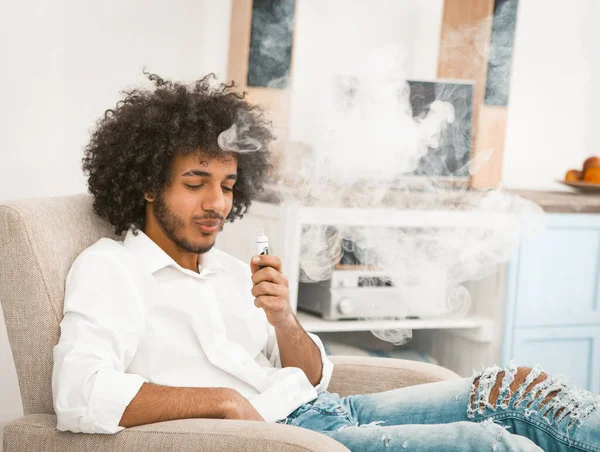Ein bärtiger Mann genießt den Qualm einer elektronischen Zigarette. Junger Mann in Freizeitanzügen sitzt zu Hause im Sessel und raucht eine elektronische Zigarette — Stockfoto
