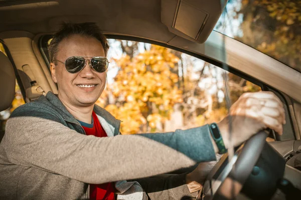 Driver stopped car in autumn forest. Handsome man in sunglasses smiles at the camera sitting in the car against the background of trees with yellow foliage outside the window