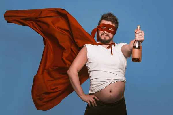 Суперовий супергерой або антигерой тримає пляшку вина, людина в супергеройській червоній масці з літаючим плащем, що тримає пляшку алкоголю, показуючи великий животик, ізольований на зів'ялому джинсовому синьому фоні — стокове фото