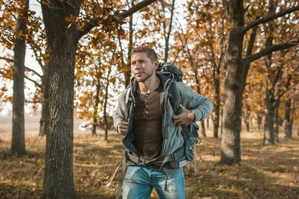 Toerist aan het begin van de reis. Een geïnspireerde man met een grote rugzak stapte uit een rode auto en begint zijn reis door het herfstbos. Wandelconcept — Stockfoto