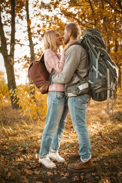 Couple de touristes amoureux dans la forêt d'automne. Jeune homme et femme baisers et câlins tout en se tenant debout sur la toile de fond de la nature d'automne coloré rétroéclairé par la lumière du soleil. Portrait intégral — Photo