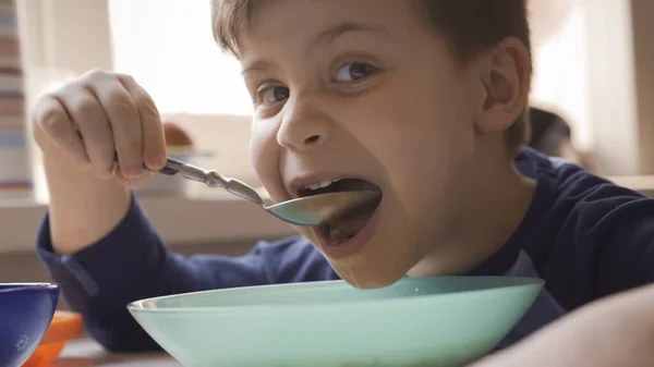 Голодный мальчик с удовольствием ест суп, глядя в камеру. Дошкольник ест себя сидя за кухонным столом с миской на нем — стоковое фото