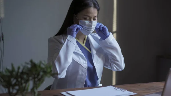 A médica trabalha com um computador no escritório dele. Mulher asiática bonita em uniforme ajusta máscara protetora enquanto sentado em uma mesa com espaços em branco nele — Fotografia de Stock