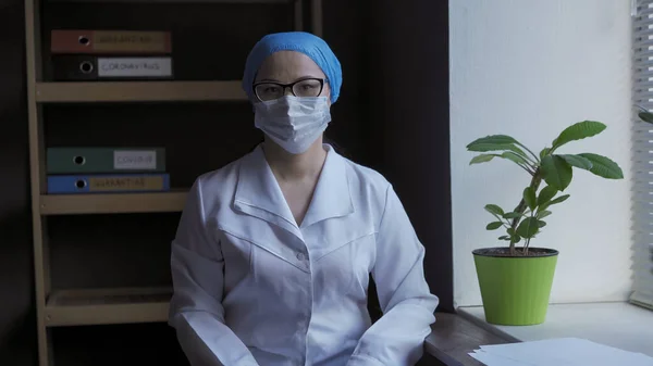 O femeie din Asia stă pe fundalul cabinetului medical. Un lucrător medical care poartă mască și haină medicală albă se uită la cameră. Conceptul de medicină și sănătate — Fotografie, imagine de stoc