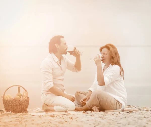 Picnic near the water. Man and woman drink red wine while sitting on a blanket on a sandy beach by the sea. Love idyll concept. Toned image — Stock Photo, Image