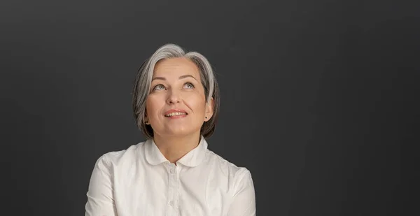 Denkende weißhaarige Geschäftsfrau, die nach oben blickt. Kreative hübsche Frau mittleren Alters lächelt nachdenklich und posiert auf grauem Hintergrund. Porträt aus nächster Nähe. Copyspace auf der rechten Seite — Stockfoto