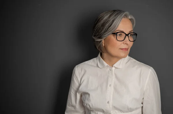 Όμορφη γκριζομάλλα επιχειρηματίας που κοιτάει στο πλάι. Ώριμη γυναίκα με γυαλιά και λευκό πουκάμισο σε γκρι φόντο. Copyspace στην αριστερή πλευρά — Φωτογραφία Αρχείου