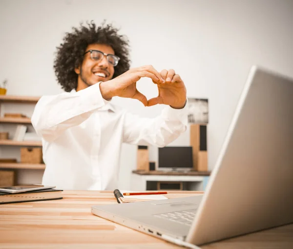 Il giovane fa videochiamate, comunica tramite collegamento video con gli amici. Un ragazzo arabo mostra un segno di cuore con le mani che esprime amore e simpatia al suo interlocutore. Immagine tonica — Foto Stock