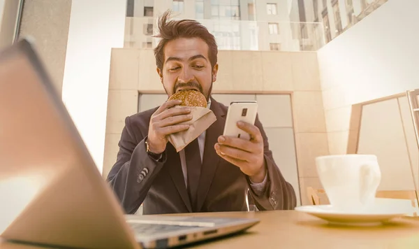 Az üzletember hamburgert eszik mobilon, miközben asztalnál ül laptoppal és egy csésze kávéval. Üzlet útközben. Gyors kaja koncepció. Hangos kép. Alsó nézet — Stock Fotó