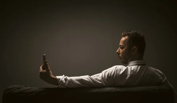 Sad businessman using mobile phone. Lonely man holds an online meeting or takes selfie with smart phone. Rear view of businessman sitting at sofa in dark interior. Copy space for text at left side — Stock Photo, Image
