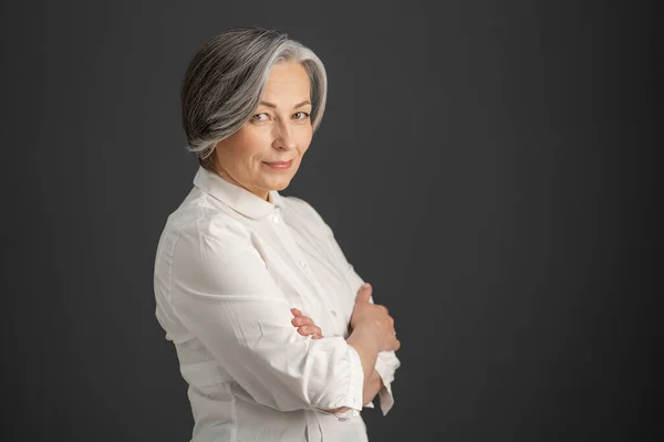 Una mujer de pelo blanco sonríe mirando a la cámara con los brazos cruzados. Elegante mujer de negocios caucásica aislada sobre fondo gris oscuro. Textspace en el lado derecho. Concepto empresarial —  Fotos de Stock