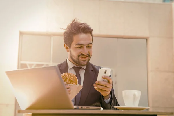 Businessman working on the go. Young man in suit eating hamburger and chatting in mobile phone while sitting at table with laptop and cup of coffee on it. Business on the go concept. Toned image
