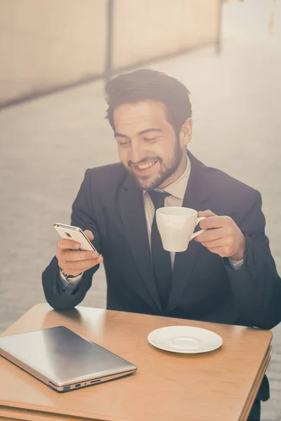 L'uomo d'affari sorridente tiene la tazza di caffè che chiacchiera in un telefono cellulare mentre si siede a un tavolo di caffè urbano all'aperto. Affari in movimento. Concetto freelance. Immagine tonica — Foto Stock