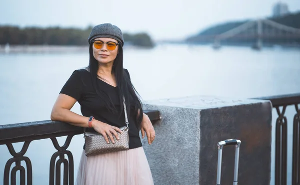 Viajera en gafas de sol descansando sobre terraplén del río. Hermosa mujer asiática con maleta viaja en la ciudad europea. Concepto de viaje —  Fotos de Stock