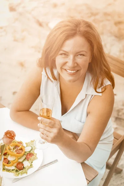 Leende kvinna håller glas vitt vin sittande vid bordet på sandstranden utomhus. Glad medelålders kvinna har datum nära havet. Hög vinkel vy. Tonad bild — Stockfoto