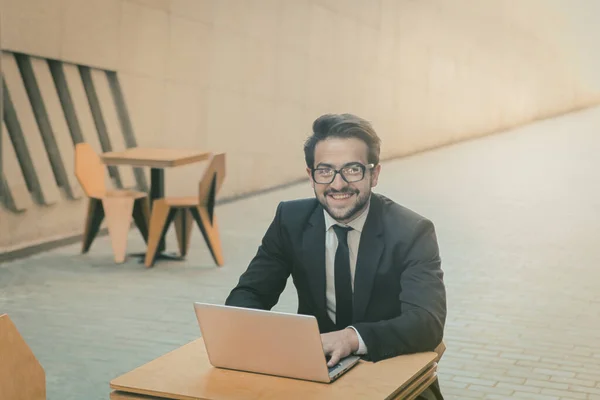 Бізнесмен працює ноутбуком у міському кафе на відкритому повітрі. Усміхнений білий чоловік дивиться на камеру за допомогою комп'ютера. Бізнес на концепції ходу. Концепція фрілансу. Тоноване зображення — стокове фото
