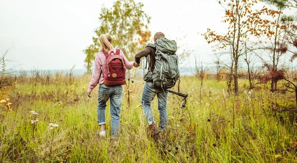 Achteraanzicht van een reizend paar backpackers die buiten door een herfstveld waden, hand in hand met wandelstokken. Wandelconcept — Stockfoto