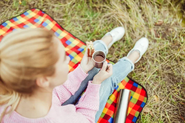 Urocza blondynka je kanapki i pije herbatę siedząc na kocu piknikowym w trawie. Skup się na kobiecych dłoniach. Strzał z bliska, widok z dużej odległości. Przekąski w naturze na świeżym powietrzu koncepcja — Zdjęcie stockowe