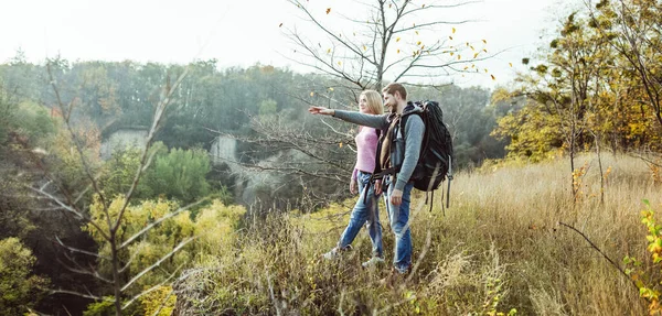 Turistas visitando los alrededores desde la cima de la colina. Hombre caucásico con mochila señala su mano en la distancia, encantadora rubia mira cuidadosamente. Viajar a pie concepto —  Fotos de Stock