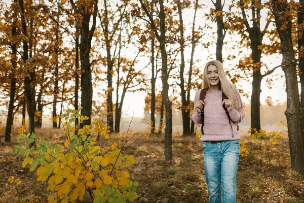 Ragazza in viaggio con uno zaino sorride in piedi su un bellissimo sfondo foresta autunnale. La bionda caucasica viaggia sulla natura in una giornata di sole. concetto di escursionismo — Foto Stock