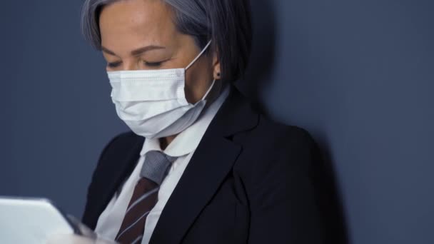 Una donna d'affari dai capelli grigi con una maschera protettiva tiene una tavoletta mentre si trova contro un muro scuro in abito da lavoro nero. Affari durante il periodo di quarantena pandemica. Concetto aziendale. Prore 422 — Video Stock