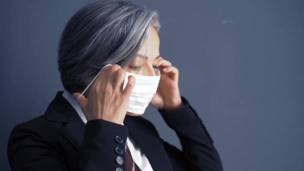 En gråhårig affärskvinna sätter på sig en skyddande mask när hon står mot en mörk vägg i en kostym. Affärer under den pandemiska karantänperioden. Affärsidé. Prores 422 — Stockvideo