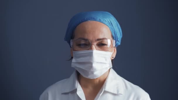 Μεγέθυνση κάμερας πάνω από τη γιατρό γυναίκα σε προστατευτική μάσκα και γυαλιά φορώντας ιατρική στολή απομονώνονται σε σκούρο μπλε φόντο. Ιατρική έννοια. Πρότυπα 422 — Αρχείο Βίντεο