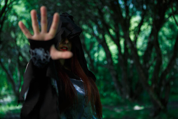 Witch with outstretched hand in black hood at dark forest