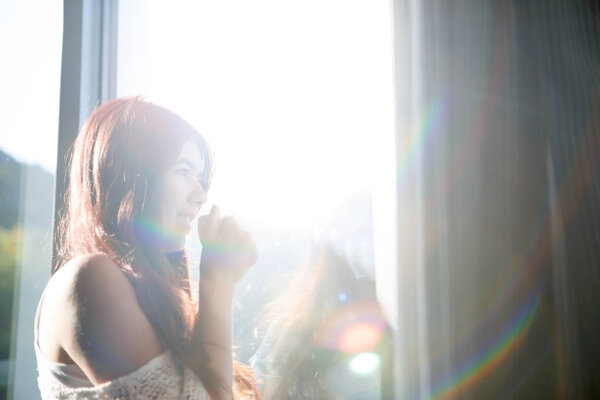 Long-haired pensive girl with cup of tea by window in room.