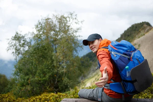 Image of sporty smiling man stretching out hand with backpack against backdrop of mountain — Stock Photo, Image