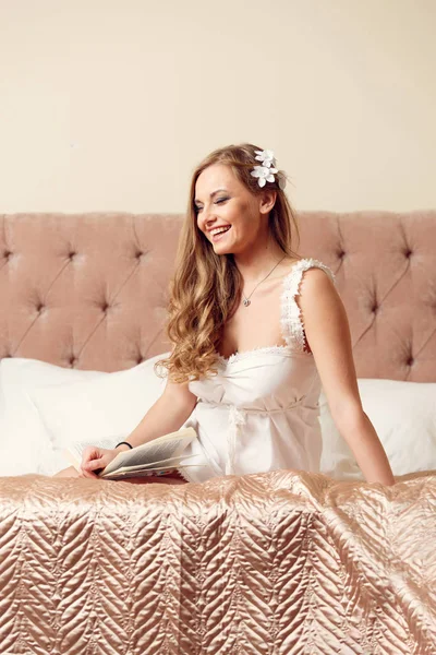 Bild einer glücklichen Frau in weißem Negligé, die auf dem Bett sitzt — Stockfoto