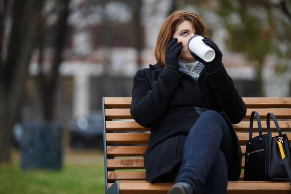 Fotka ženy v černém kabátě, mluví o telefonu a pití kávy sezení — Stock fotografie