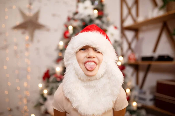 Foto de niña en el sombrero de Santa con barba mostrando la lengua contra el fondo de las decoraciones de Año Nuevo —  Fotos de Stock