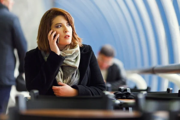 Imagen de una chica de abrigo negro hablando por teléfono sentada en la cafetería —  Fotos de Stock