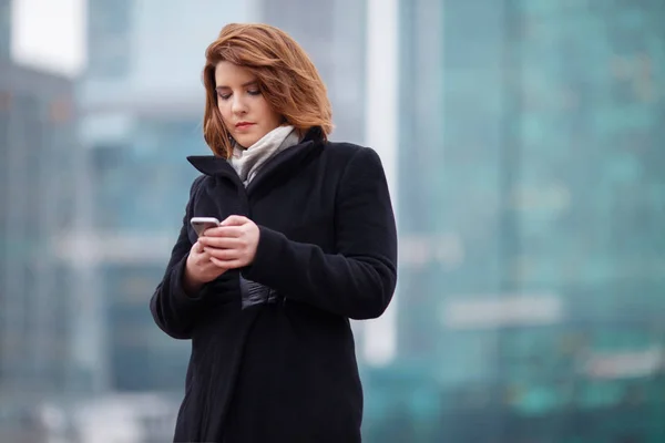 Изображение девушки в черном пальто с телефоном в руках на фоне современного здания — стоковое фото