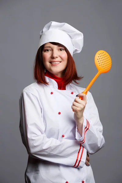 Foto cocinero morena en bata blanca y gorra con cuchara ranurada — Foto de Stock