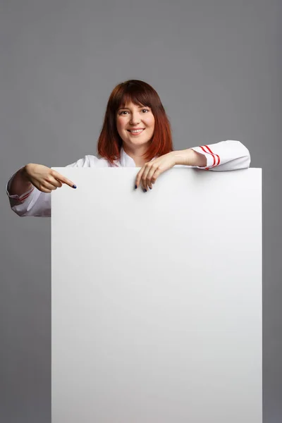 Foto de cozinheira mulher sorridente em roupão branco apontando o dedo para a folha vazia para inscrição — Fotografia de Stock