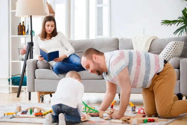 Family photo of father and son playing on floor in toy road on background of pregnant woman reading book lying on couch — Stock Photo, Image