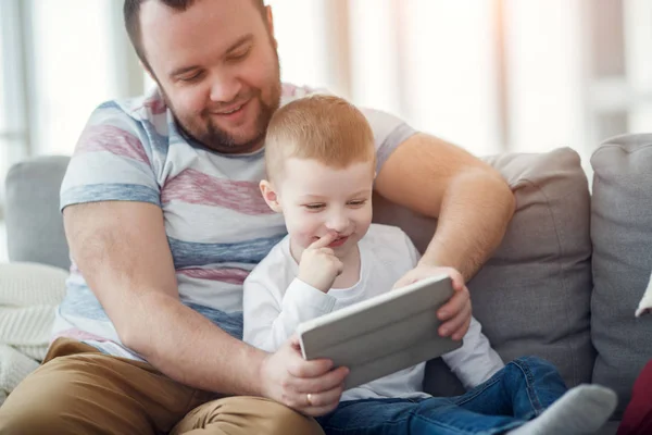 Foto do pai com seu filho com tablet sentado no sofá cinza — Fotografia de Stock