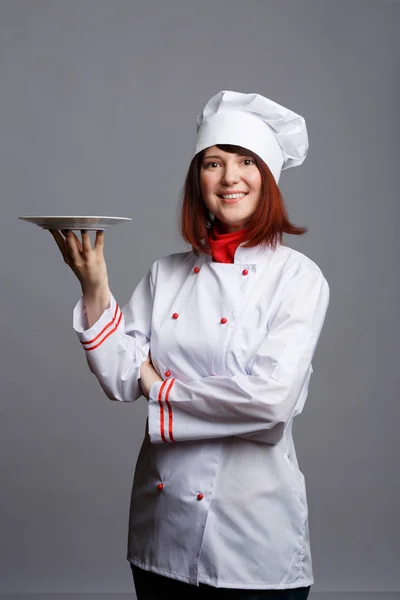 Φωτογραφία χαμογελώντας σεφ κορίτσι σε λευκό παλτό με ένα άδειο πιάτο — Φωτογραφία Αρχείου