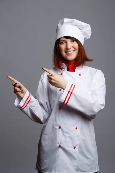 Φωτογραφία χαμογελαστό κορίτσι σεφ σε λευκή ρόμπα επισημαίνοντας δάχτυλα στο κενό χώρο — Φωτογραφία Αρχείου