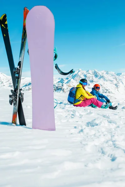 Foto de snowboard, esquís en el fondo de la mujer deportiva sentada y el hombre en la colina nevada —  Fotos de Stock